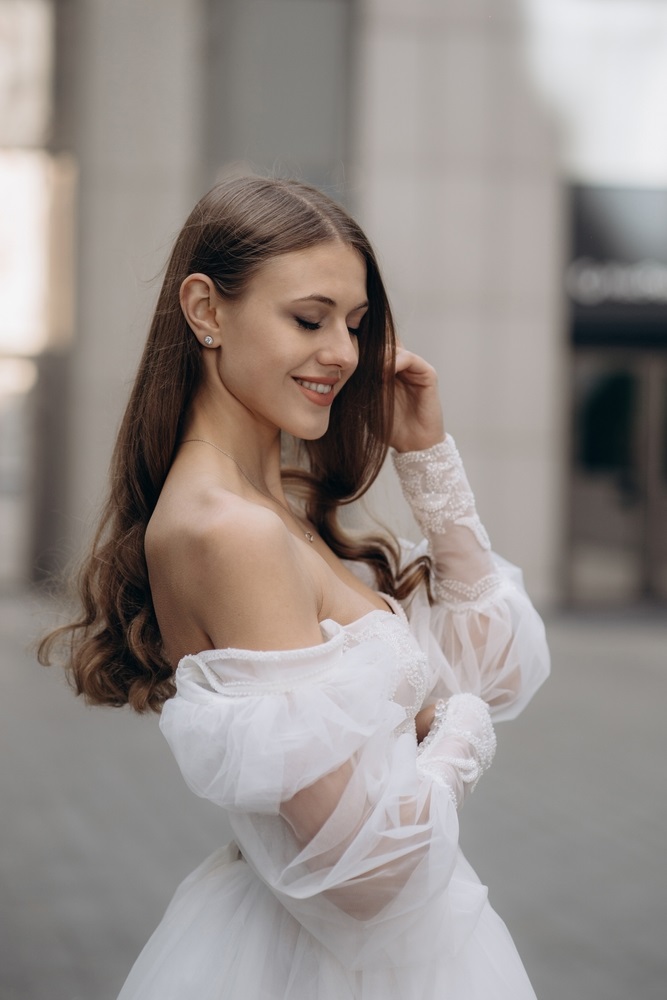 Alt text: Portrait of beautiful bride with long hair in white wedding dress. Fashionable long sleeves white wedding dress with bare shoulders, beads, lace, and neckline.